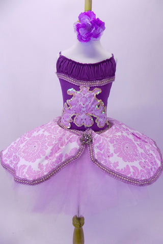 White pull on English-style tutu has layers of crisp white tulle with layers of soft  lavender tulle on top, The figure-8 overlay is a brocade pattern in lavender and white with corded trim and large broach accent at front and back. The separate purple leotard has gathered front accent and matching sequin lined brocade inserts at front and back-sides. Comes with matching floral hair accessory. Front