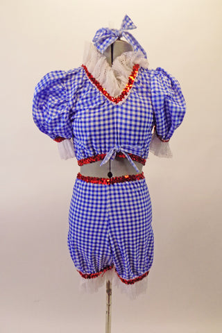 Two-piece blue and white gingham ragdoll themed costume has bloomer style bottom pouffe sleeved half top. There is a wide white lace ruffle at legs, sleeves and neckline with edged with a red sequin accent. Comes with hair bow accessory. Front