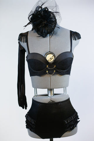 Black bustier style bra with fringed epaulets and gold chain details. at front. Has ruffled, high-waisted panty, long black gloves and a black head piece. Front zoom