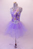 Lavender romantic tutu has a glitter tulle skirt with large curly hem. The bodice has gold and purple sequined accents with a single right and triple left shoulder strap that form a unique angle at the back. There is a large lavender flower accent at the right shoulder and left hip. Comes with a gold hair barrette. Left side