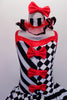 Harlequin themed tutu dress is an alternating pattern of black-white stripes & black-white checks. A red tulle skirt with black satin ribbon makes the skirt & matches the ruffled collar. Three red bows accent the bodice. Comes with red hair bow & stirrup socks. Front zoomed
