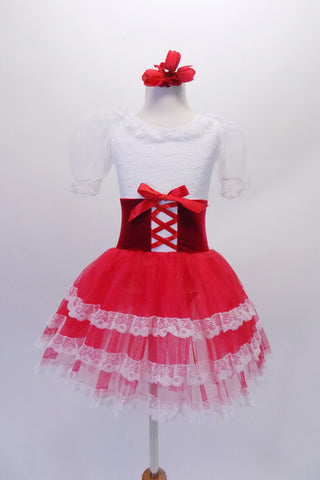 Tutu dress has a white lace bodice with lace neck ruffle and sheer pouffe sleeves. The red velvet wide waistband has a delicate lace-up tie accent at the front. The red triple-layer tulle skirt has a wide lace edging. Comes with a floral hair accessory. Front