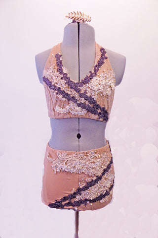 Nude based 2-piece costume has a halter neck bra top and brief style shorts. White breaded and crystalled bridal applique adorns both top and bottom which are further accented with narrow, angled bands of embroidered lace applique in a purple/grey hue. Comes with hair barrette. Front