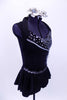 Black short unitard has peplum skirt & shawl collar. Bodice has mesh-chain design, silver cording & crystal accents. Has large beaded broach & hair accessory. Right side
