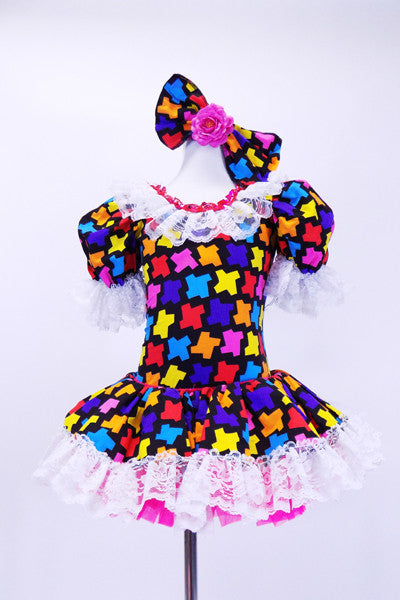 Black leotard dress has colorful puzzle piece decorative front with lace trim at neck sleeves and skirt. Dress has bright pink petticoat and matching bow hair accessory. Front