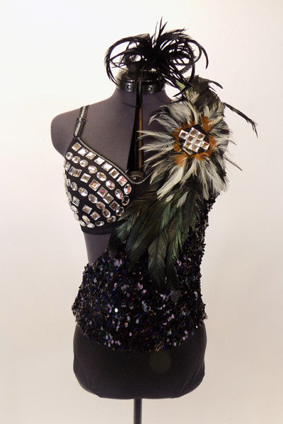 Black bra is covered with large jewel accents & sits beneath one shoulder, black sequined leotard dress with large feather accent on shoulder. Has feather hair accessory. Front.