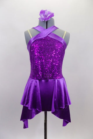 Purple sequined bodice is accented by wide satin banding that crosses over across the chest. The attached satin skirt i falls over the hips and is longer at the two sides. Comes with floral hair accessory. Front