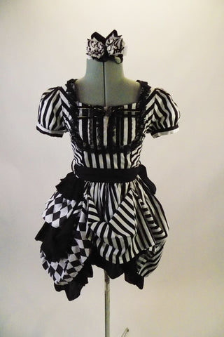 Black and white striped saloon girl themed dress has a laced bib bust area and short pouffe sleeves. The right hip is gathered with pick-ups in layers of black and black/white checkers. The back of the dress has lace-up detail. Comes with matching hair accessory. Front