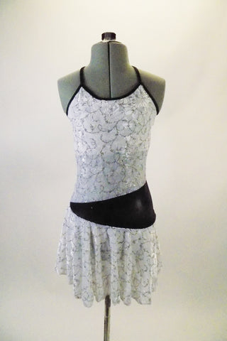 White knee-length dress has silver swirl pattern and black piping with back cross straps. The black waistband starts at the right hip and widens as it wraps around the back and front joining at the left side. Comes with a hair accessory. Front