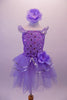 Lavender ballet dress has a rose ribbon bodice with floral sequin accents. The soft tulle layered skirt is accented with silver sequin-edged double peplum and a matching shoulder ruffle. A large lavender peony flower graces the left hip. Comes with matching peony floral hair accessory. Front