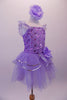 Lavender ballet dress has a rose ribbon bodice with floral sequin accents. The soft tulle layered skirt is accented with silver sequin-edged double peplum and a matching shoulder ruffle. A large lavender peony flower graces the left hip. Comes with matching peony floral hair accessory. Right side