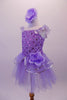 Lavender ballet dress has a rose ribbon bodice with floral sequin accents. The soft tulle layered skirt is accented with silver sequin-edged double peplum and a matching shoulder ruffle. A large lavender peony flower graces the left hip. Comes with matching peony floral hair accessory. Left side
