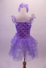 Lavender ballet dress has a rose ribbon bodice with floral sequin accents. The soft tulle layered skirt is accented with silver sequin-edged double peplum and a matching shoulder ruffle. A large lavender peony flower graces the left hip. Comes with matching peony floral hair accessory. Back