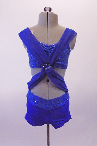 Royal blue costume is a sequined & crystal lines bra top and booty shorts. Two wide chiffon kerchief like bands originate from the back straps and cross over at the front. A large jewelled brooch attached the bands together at the base of the bra front, from where they extend out & are attached at the hips. Front