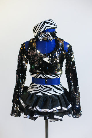 Black sequined top has long sleeves, high neck with zebra band and &bow. Open back has corset ties & is complimented by a ruffled black skirt & zebra pill hat. Front